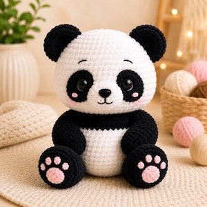 May include: A handmade crochet panda plush toy featuring a white head and torso with black ears, arms, and legs. The panda has large black eyes, a small black nose, and light pink blush marks on its cheeks. Its feet display pink paw pads.
