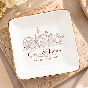Puede incluir: Un plato cuadrado de cerámica blanca con borde dorado. El centro presenta una ilustración lineal en marrón del horizonte de Las Vegas, el texto "Las Vegas", los nombres "Olivia & James" y la fecha "09.21.25".