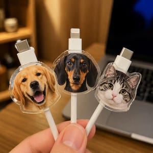 May include: Three white charging cables featuring clear acrylic charms attached to the connectors. The charms display high-resolution portraits of a golden retriever, a black and tan dachshund, and a grey and white tabby cat face.