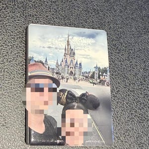 Könnte beinhalten: Ein rechteckiger Magnet mit einem aufgedruckten Foto eines großen Schlosses mit hohen Türmen und blauen Dächern. Das Bild zeigt eine Menschenmenge vor dem Schloss, mit zwei Personen im Vordergrund, die Hüte und Ohren tragen.