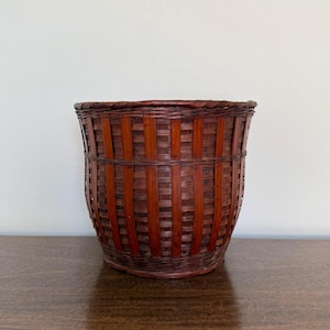 May include: A cylindrical basket crafted from dark reddish-brown woven bamboo strips. The structure features vertical slats reinforced by horizontal bands, creating a sturdy, open-weave pattern with a solid, flat base and a rounded rim.