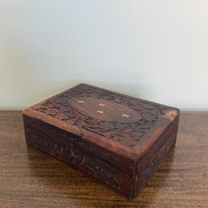 May include: A rectangular dark brown wooden box featuring intricate floral carvings across the lid and sides. The centre of the lid displays an oval inlay with small, light-coloured leaf-shaped brass accents embedded into the wood surface.