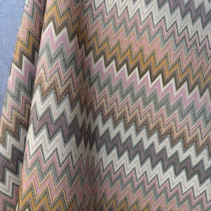 Könnte beinhalten: Ein Textilstoff mit einem sich wiederholenden Zickzackmuster in den Farben Rosa, Senfgelb, Grau und Weiß. Das Material hat eine strukturierte, gewebte Optik mit vertikalen Falten, die Tiefe und Schatten über das Zickzack-Design erzeugen.