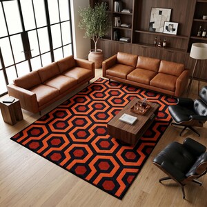 Op de afbeelding: Een rechthoekig vloerkleed met een herhalend geometrisch patroon van in elkaar grijpende oranje en rode zeshoeken op een zwarte achtergrond. Het kleed ligt in een woonkamer met twee bruine leren banken, twee zwarte fauteuils en een houten salontafel.