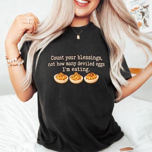 Peut inclure: Un t-shirt noir avec le texte "Count your blessings, not how many deviled eggs I'm eating." centré au-dessus d'une illustration de trois œufs farcis avec des jaunes jaunes et une garniture verte, imprimé en beige clair.