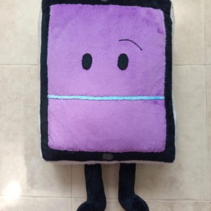 Puede incluir: Un juguete de peluche con forma de tableta rectangular con pantalla morada, borde negro y dos piernas negras. La pantalla presenta dos ojos ovalados negros, una ceja negra curva y una línea horizontal de color azul claro en el centro.