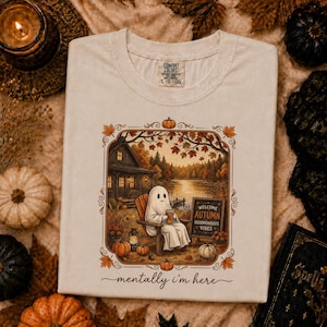 Könnte beinhalten: Ein cremefarbenes T-Shirt mit einer Grafik eines Geistes, der in einem Holzstuhl an einem See sitzt und eine Tasse hält. Das Motiv zeigt eine Hütte, Kürbisse und ein Schild mit der Aufschrift "WELCOME AUTUMN HARMONIOUS VIBES". Unten steht "mentally i'm here".
