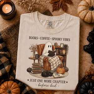 Könnte beinhalten: Ein cremefarbenes T-Shirt mit einer Grafik eines Geistes, der in einem Sessel sitzt und ein Buch liest. Der Text lautet: BOOKS • COFFEE • SPOOKY VIBES und JUST ONE MORE CHAPTER before bed. Die Illustration zeigt ein Bücherregal und Kürbisse.