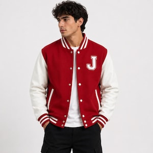 Puede incluir: Una chaqueta universitaria roja con mangas blancas y un parche con la letra J blanca en el pecho izquierdo. La chaqueta cuenta con cierre frontal de botones a presión, cuello acanalado y puños a rayas. Se lleva sobre una camiseta blanca.