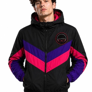 May include: A black hooded jacket featuring a vibrant chevron pattern across the chest in purple and hot pink. The jacket has a full-length front zipper, ribbed cuffs, and a circular patch on the left chest displaying a stylized face graphic.