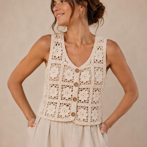 May include: A sleeveless cream-colored crochet vest featuring a repeating square floral pattern. The garment has a V-neckline and a front button closure with four round, light brown wooden buttons. The crochet work displays an intricate openwork design.