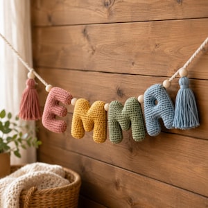 May include: A decorative garland featuring five crocheted letters spelling the name EMMA in muted tones of dusty rose, mustard yellow, sage green, and slate blue, accented with wooden beads and two yarn tassels on a thick white twisted rope.