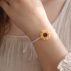 May include: A delicate bracelet featuring a small, crocheted sunflower charm with yellow petals and a dark brown center, attached to a thin, off-white cord. The bracelet has adjustable ends with small knots, showcasing a simple, handcrafted design.