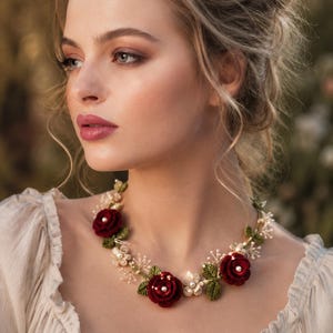 May include: A floral necklace featuring deep red crocheted roses, small white crocheted flowers, and green crocheted leaves. The design is accented with small white pearls and delicate sprigs, all arranged on a thin, flexible wire base.