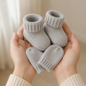 May include: A pair of light grey knitted baby booties and a matching pair of light grey knitted baby mittens. The booties feature a folded ribbed cuff, and the mittens have a ribbed wrist band, both crafted from a soft, solid-coloured yarn.