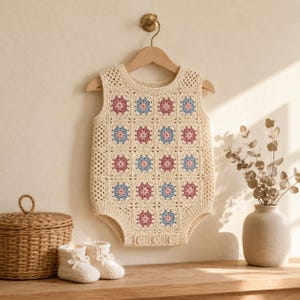 May include: A sleeveless crochet romper featuring a cream-colored base with a repeating pattern of square motifs in dusty pink and light blue. The garment has a round neckline, a textured stitch design, and three small buttons at the bottom hem.