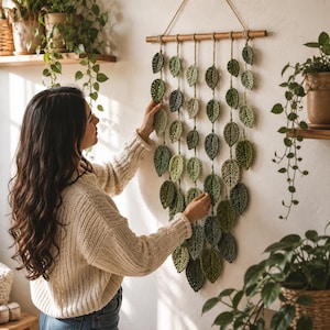 May include: A wall hanging featuring multiple strands of crocheted leaves in various shades of green, suspended from a horizontal wooden dowel. The leaves are crafted with textured yarn and hang at different lengths, creating a cascading effect.