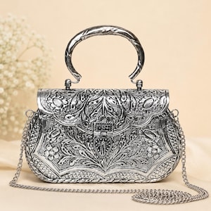 Puede incluir: Un bolso de mano de metal plateado con un intrincado patrón floral y de hojas en relieve en la solapa frontal y el cuerpo. El bolso incluye un asa superior curva, un cierre central en forma de corazón y una fina correa de cadena plateada.