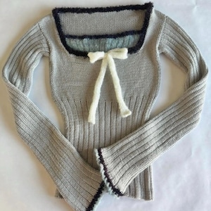May include: A long-sleeved, light grey ribbed knit jumper featuring a square neckline trimmed with dark navy blue yarn. A white knitted bow is attached at the centre of the neckline. The sleeve cuffs also feature a dark navy blue trim.