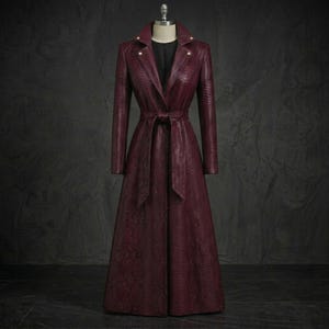 May include: A long, deep burgundy trench coat featuring a textured snakeskin pattern throughout. The coat includes a notched lapel, long sleeves, a matching tie belt at the waist, and a black inner layer visible at the neckline, displayed on a mannequin.