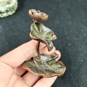 May include: A small bronze-toned metal sculpture featuring a frog resting on a large lily pad with a tall stem supporting a blooming lotus flower above. The piece has a weathered, dark metallic finish with visible textures on the leaves and petals.