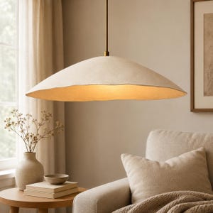 Może przedstawiać: Lampa wisząca z szerokim, płytkim kloszem w kształcie kopuły w kolorze złamanej bieli, o teksturowanej, organicznej krawędzi. Klosz zawieszony jest na cienkim, prostym, mosiężnym pręcie, emitując ciepłe, delikatne światło od spodu kopuły.