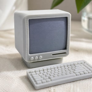 May include: A miniature light gray computer model featuring a boxy monitor with a dark gray screen and three circular buttons below it. A matching light gray rectangular keyboard with raised keys sits in front of the monitor on a flat surface.