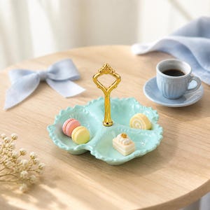 May include: A light mint green three-section ceramic serving dish with a central gold-colored metal handle. The dish contains miniature food items including a pink macaron, a yellow macaron, a small cake slice, and a tiny rolled sponge cake.