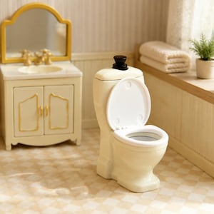 Könnte beinhalten: Eine cremefarbene Miniatur-Toilette mit weißem Sitz und Deckel sowie einem schwarzen Spülknopf auf dem Spülkasten. Die Toilette ist in einem Puppenhaus-Badezimmer neben einem passenden cremefarbenen Waschtisch mit goldfarbenem Spiegel platziert.