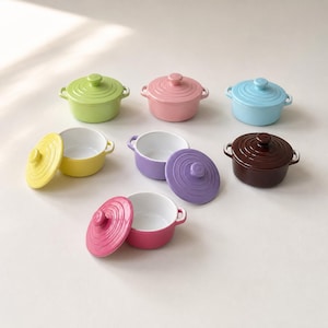 Könnte beinhalten: Eine Sammlung von sieben kleinen, runden Keramiktöpfen mit passenden Deckeln in verschiedenen Farben wie Limettengrün, Pastellrosa, Hellblau, Gelb, Lavendel, Dunkelbraun und Magenta. Jeder Topf hat zwei seitliche Griffe und ein weißes Inneres.
