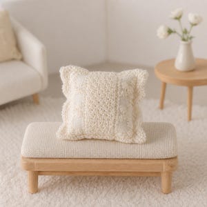 May include: A square, cream-coloured crocheted throw pillow with a textured, intricate stitch pattern and ruffled edges. The pillow is resting on a rectangular wooden bench with a light-coloured, woven fabric seat and four short, sturdy wooden legs.