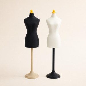 May include: Two miniature dress forms standing side by side. One form is black with a beige stand, and the other is white with a black stand. Both forms feature a small yellow knob at the neck, providing a clear view of their upright structure.