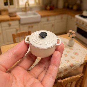 May include: A miniature off-white round Dutch oven pot with a matching lid and a small black knob handle. The pot features two side handles and a circular ridged design on the lid, presented as a small-scale kitchen accessory for dollhouses.