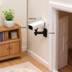 May include: A miniature white security camera with a black mounting bracket attached to a wall. The camera measures 3cm/1.2in in length, 1cm/0.4in in height, and the bracket measures 2cm/0.8in in height as indicated by the printed text labels.