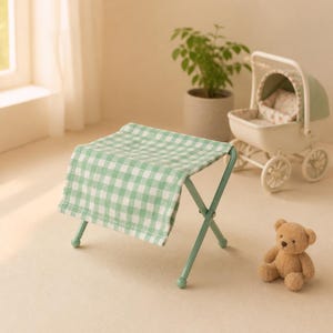Puede incluir: Un taburete plegable pequeño con un asiento de tela de cuadros vichy en verde claro y blanco, y una estructura de metal verde claro a juego. El taburete presenta un diseño de patas en cruz con cuatro pies redondeados para una base estable.