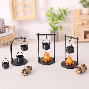 May include: A set of miniature black metal campfire stands with hanging pots, two featuring orange and yellow flame accents on circular bases, accompanied by small bundles of brown wooden logs tied with twine and a separate small black pot.