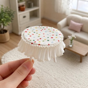 Puede incluir: Una mesa redonda en miniatura con una superficie blanca decorada con un patrón de lunares coloridos en rojo, verde, amarillo y azul. La mesa está cubierta con una falda de tela blanca con borde de volantes y pequeñas patas blancas.