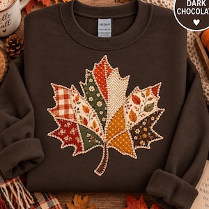 May include: A dark chocolate brown crewneck sweatshirt featuring a large, central maple leaf graphic composed of various autumn-themed fabric patches in shades of orange, red, green, and cream, all outlined with decorative white stitching.