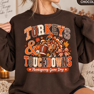May include: A dark chocolate brown sweatshirt featuring a graphic print with the text "TURKEYS & TOUCHDOWNS" and "Thanksgiving Game Day". The design includes a turkey wearing a beanie, scarf, and sunglasses, holding a football, with pumpkins.