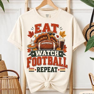 May include: An ivory short-sleeve t-shirt featuring a graphic of a turkey wearing sunglasses, a football, a pumpkin pie, and a glass of beer. The text reads EAT WATCH FOOTBALL REPEAT in red and green block letters, surrounded by autumn maple leaves.