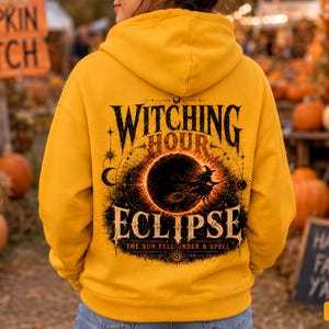 May include: A gold hooded sweatshirt featuring a black graphic on the back. The design displays a silhouette of a witch on a broomstick flying in front of a large moon, with the text Witching Hour Eclipse and The Sun Fell Under A Spell.