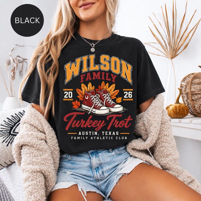 May include: A black t-shirt featuring a graphic with the text "WILSON FAMILY" in orange, "20 26" on the sides, and "Turkey Trot" in red. The design includes a pair of red high-top sneakers surrounded by orange and yellow autumn leaves.