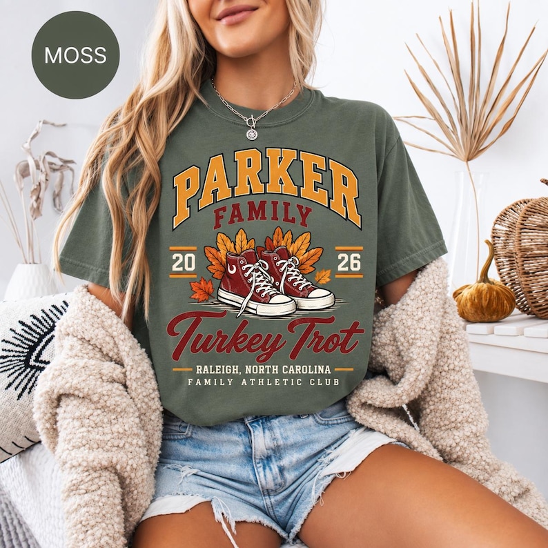 May include: A moss green t-shirt featuring a graphic with the text Parker Family Turkey Trot 2026, Raleigh, North Carolina, Family Athletic Club. The design includes a pair of red high-top sneakers surrounded by orange and yellow autumn leaves.