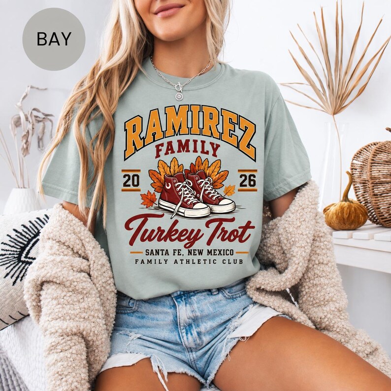 May include: A light sage green t-shirt featuring a graphic print with the text RAMIREZ FAMILY, Turkey Trot, SANTA FE, NEW MEXICO, FAMILY ATHLETIC CLUB, and the numbers 20 and 26. The design includes illustrations of autumn leaves and red sneakers.