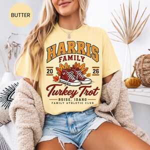 May include: A butter yellow short-sleeve t-shirt featuring a graphic with the text Harris Family Turkey Trot Boise, Idaho Family Athletic Club, 2026, and an illustration of two red high-top sneakers surrounded by orange and yellow autumn leaves.