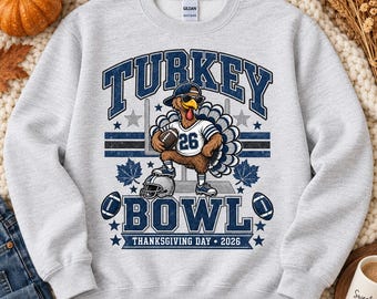 May include: An ash grey sweatshirt featuring a graphic of a turkey wearing sunglasses and a baseball cap, holding a football. The text reads TURKEY BOWL THANKSGIVING DAY 2026. The design includes football elements, maple leaves, and stars in navy blue.