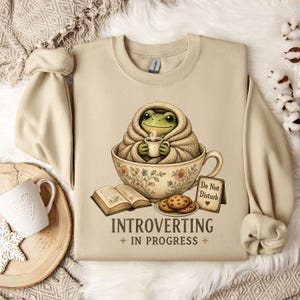 May include: A beige crewneck sweatshirt featuring a graphic of a green frog wrapped in a blanket, sitting inside a floral teacup while holding a small mug. Beside the cup are an open book, a plate of cookies, and a sign that reads Do Not Disturb. The text below says INTROVERTING IN PROGRESS.