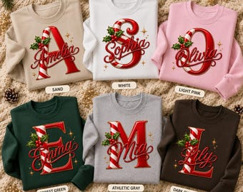 May include: Six crewneck sweatshirts in sand, white, light pink, forest green, athletic gray, and dark chocolate. Each features a large red and white candy cane letter with a holly sprig and a cursive name: Amelia, Sophia, Olivia, Emma, Mia, Lily.