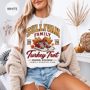 May include: A white t-shirt featuring a graphic with the text SULLIVAN FAMILY, 2026, Turkey Trot, MADISON, WISCONSIN, FAMILY ATHLETIC CLUB, and an illustration of two red sneakers surrounded by orange and yellow autumn leaves.