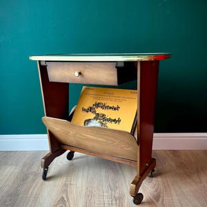 May include: A wooden side table on wheels featuring a single drawer with a small round knob and a lower magazine rack. The top surface has a gold-toned metal trim. A yellow Vivaldi vinyl record sleeve is stored in the rack below the drawer.